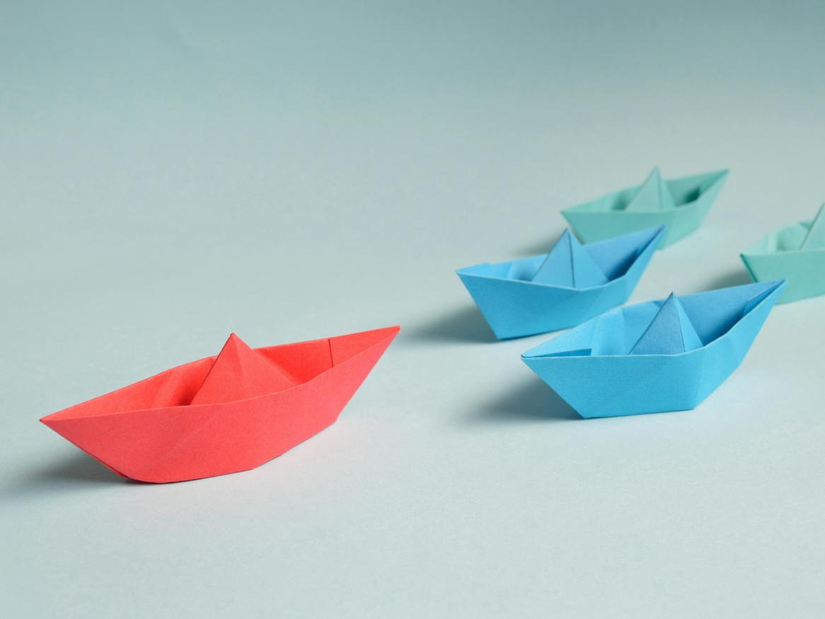 Blue paper boats against a plain background, with a red boat in the lead.